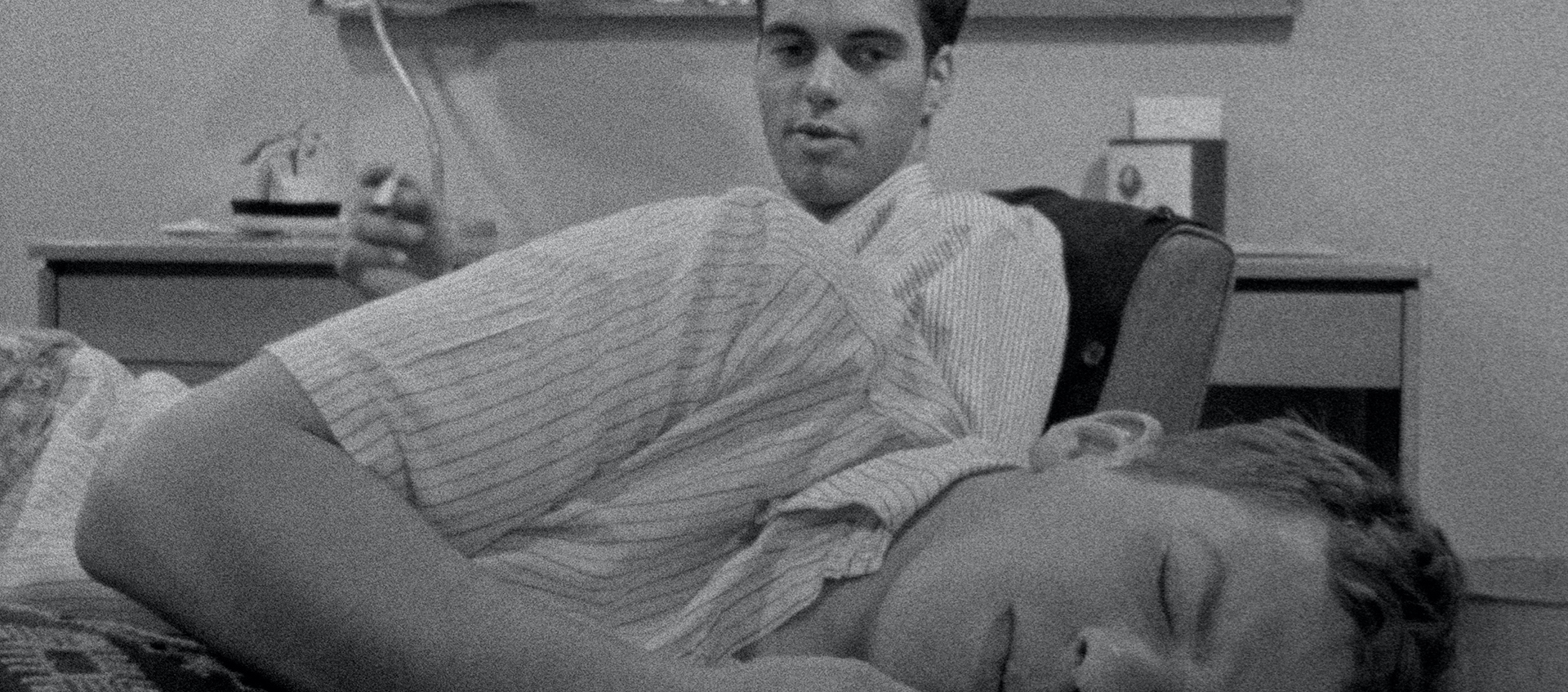 A young man is laying down and another young man is sitting behind him holding a cigarette. A desk, lamp, and bulletin board are in the background.