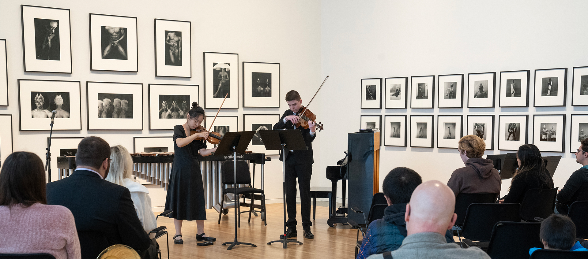 A seated audience watches as two people play violins in a gallery. The musicians stand in front of framed black-and-white photographs.