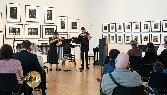 A seated audience watches as two people play violins in a gallery. The musicians stand in front of framed black-and-white photographs.