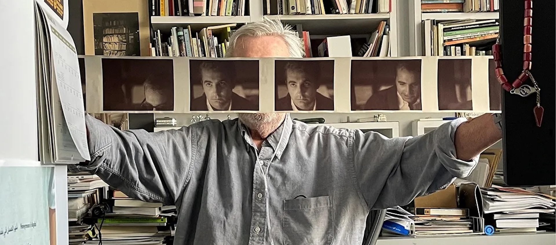 A man sits at a desk holding a horizontal printout of pictures of himself in front of his face.