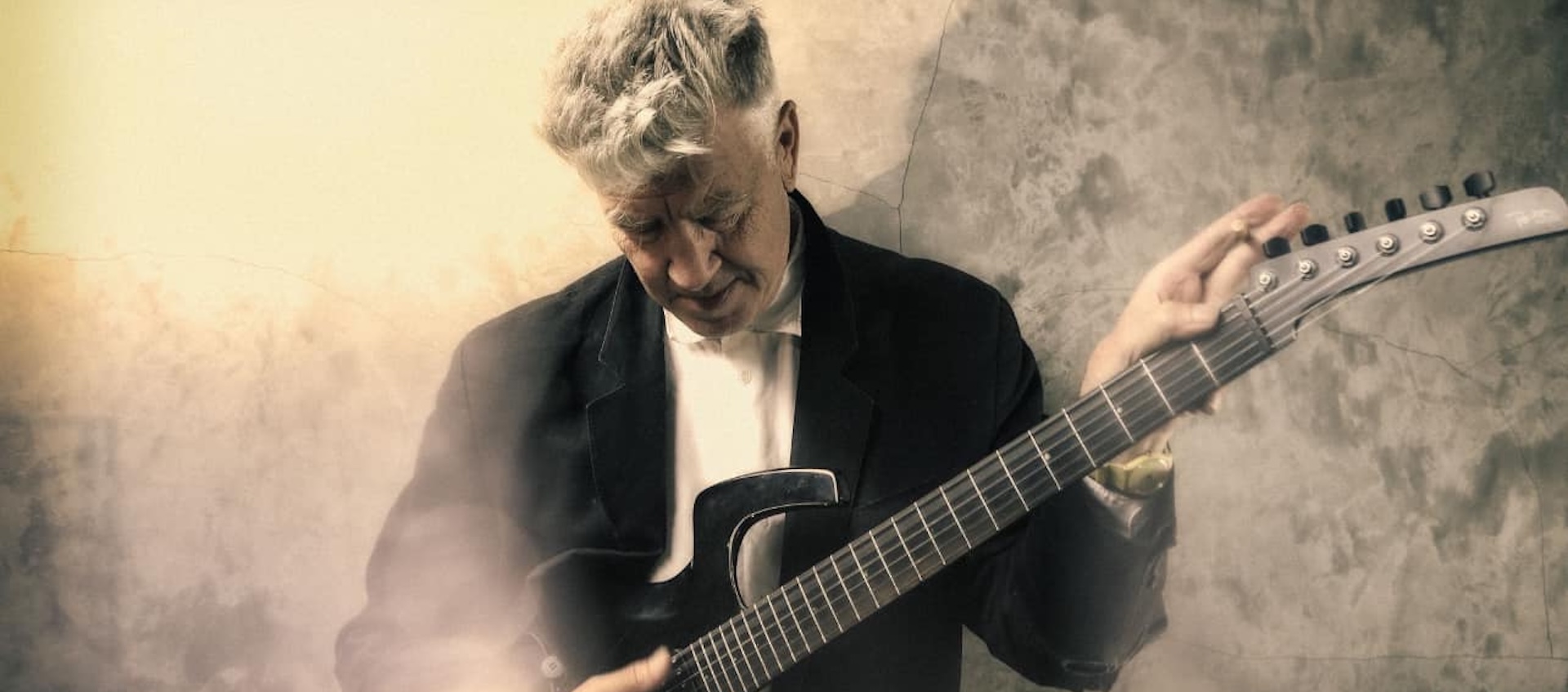 Sepia-toned photo of a white-haired man as he looks down at the electric guitar across his chest.