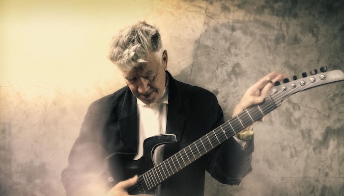 Sepia-toned photo of a white-haired man as he looks down at the electric guitar across his chest.