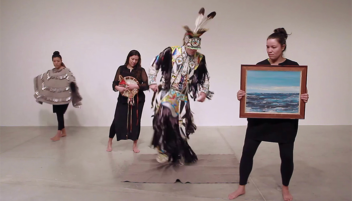 Four performers wear Indigenous ceremonial or black clothing. One wears a gray felt shawl with silver jingles. One holds a landscape painting.